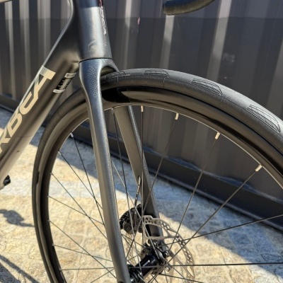 Quadro dianteiro e roda de bicicleta preta com texto ORBEA em fundo de pavimento e parede metálica.
