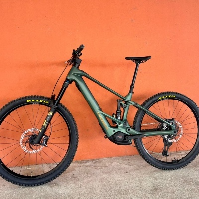 Bicicleta de montanha elétrica Orbea verde fosco com rodas Maxxis pretas e parede laranja ao fundo