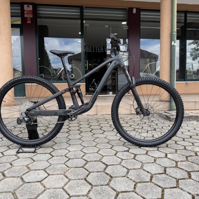 Bicicleta de montanha preta com quadro TREK em pavimento de blocos hexagonais, loja ao fundo.