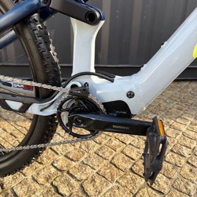 Peça de bicicleta elétrica com pedal e corrente em preto, quadro cinza-claro e azul metálico, pneu robusto.