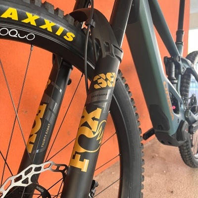 Bicicleta elétrica cinza com suspensão FOX 38, pneu MAXXIS e freio de disco junto a parede laranja