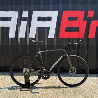 Bicicleta de corrida preta Orbea apoiada em suporte com fundo preto, texto vermelho e branco.