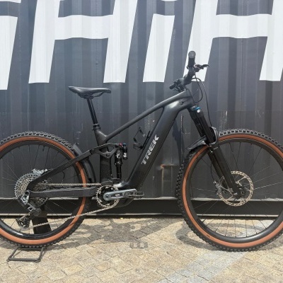 Bicicleta de montanha elétrica preta com quadro TREK apoiada num suporte de chão à frente de uma parede preta com letras brancas.