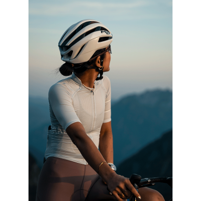 Mulher de ciclismo com capacete branco e roupa justa, segurando guidão de bicicleta em paisagem montanhosa