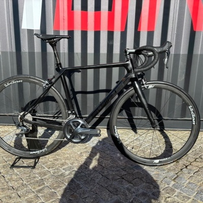 Bicicleta de estrada preta com rodas Vision e grupo Shimano Ultegra encostada a uma parede metálica.