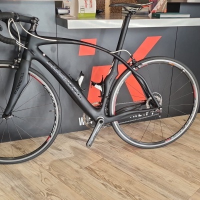 Bicicleta de estrada preta mate com detalhes vermelhos e brancos ao lado de um balcão preto com logo vermelho.