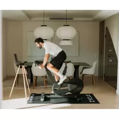 Homem a usar bicicleta estática preta no interior com mesa e cadeiras ao fundo