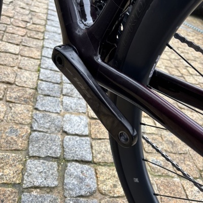 Pedalier preto de bicicleta Shimano 105 com quadro roxo escuro e roda traseira em pneu preto sobre piso de pedra