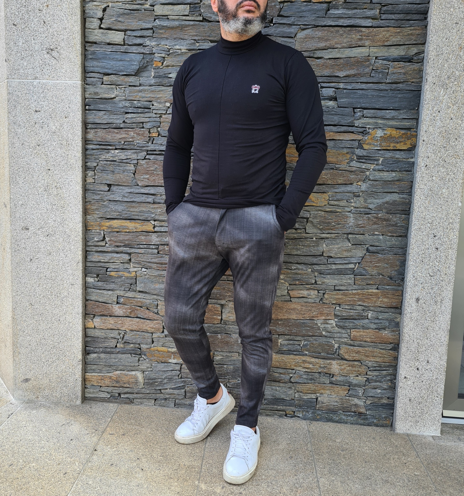 CALÇAS TECIDO SLIM FIT Homem com camisola preta e calças cinzentas em parede de pedra
