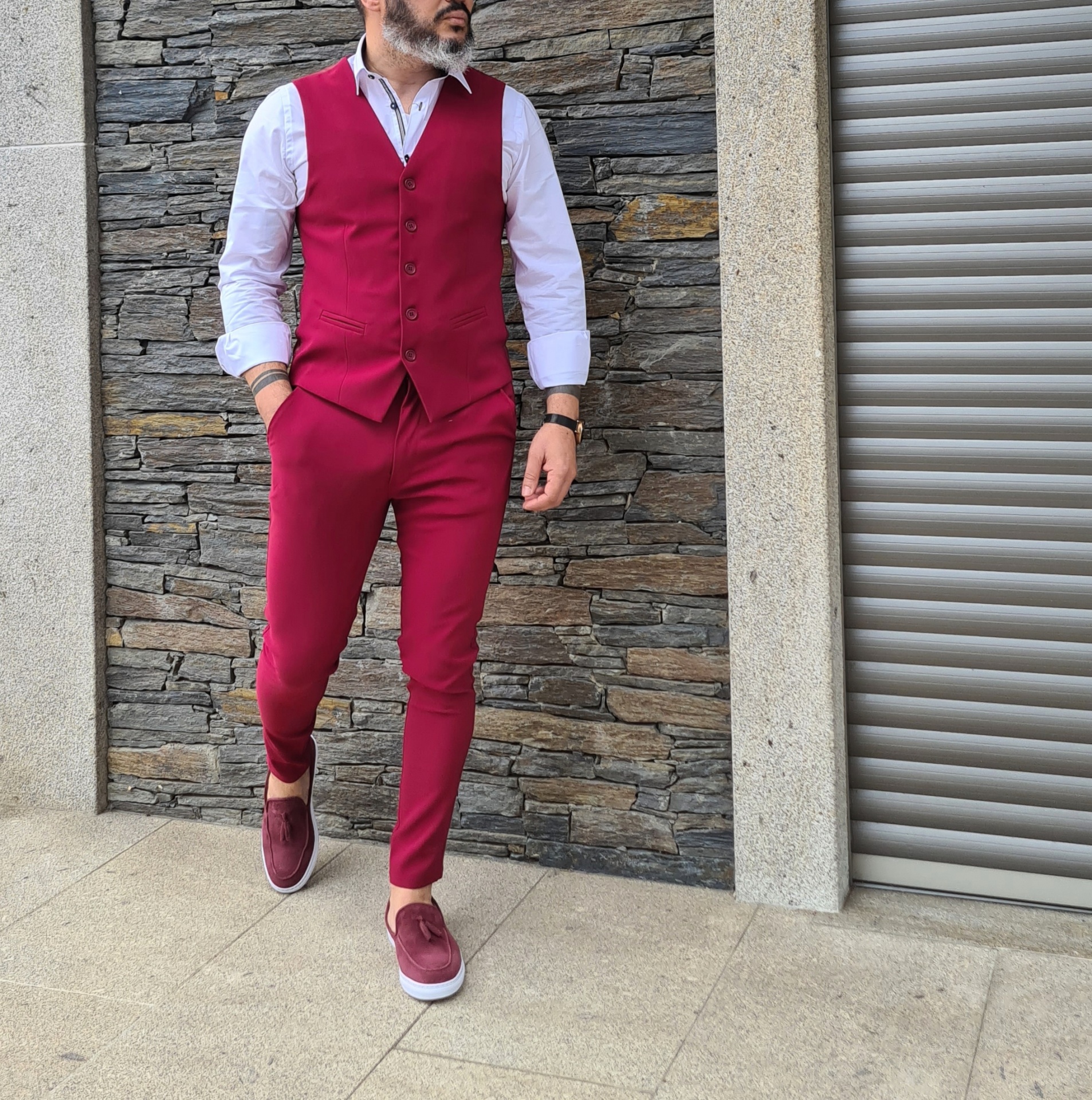 Calça cor cereja Homem usando fato vermelho com colete e calças, camisa branca e ténis vermelhos posa em frente a parede de pedra
