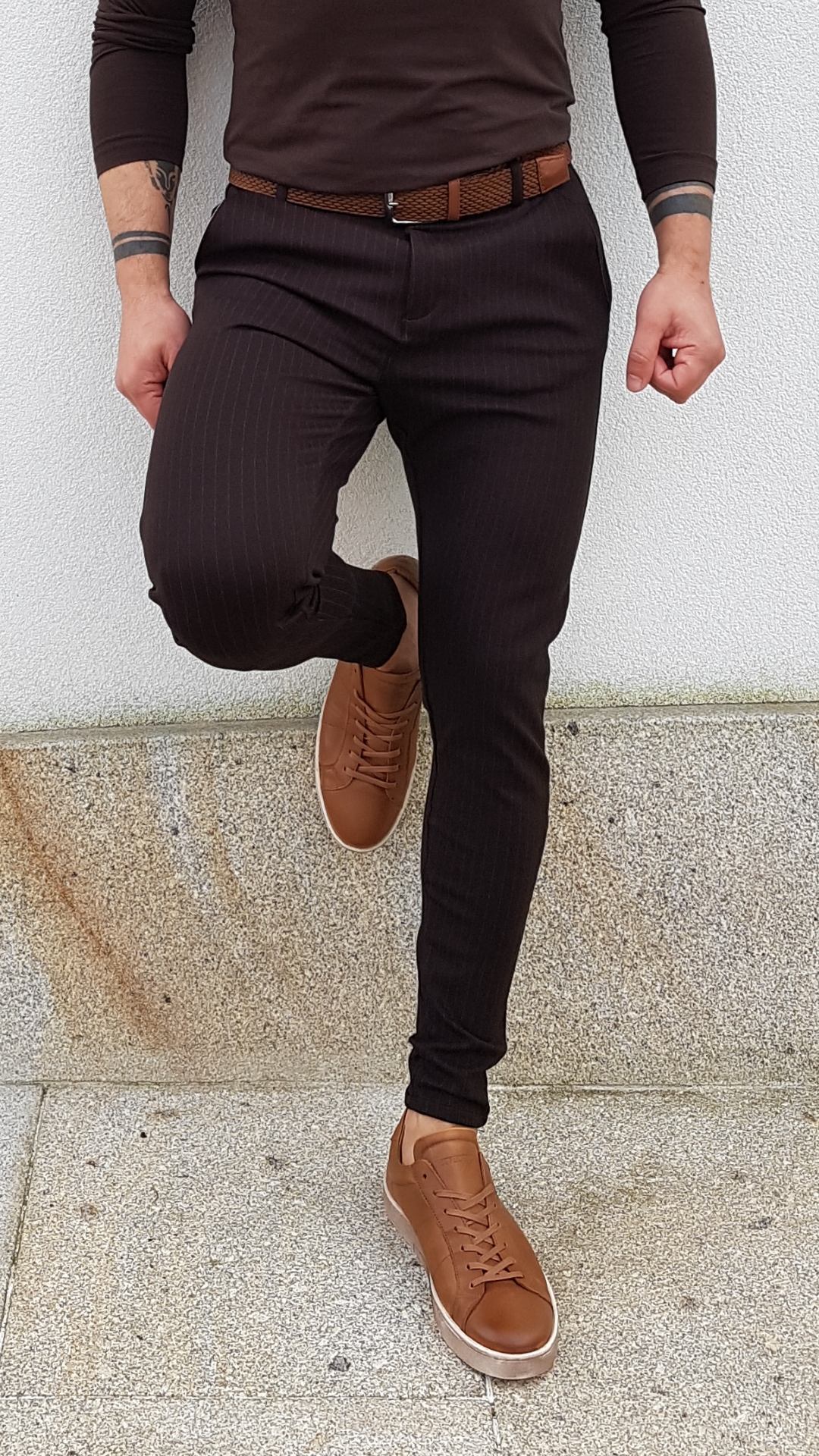 CALÇAS TECIDO SLIM FIT,CASTANHO ESCURO Homem vestido com calças pretas risca de giz, cinto castanho e sapatos castanhos, em fundo de parede branca e chão de cimento.