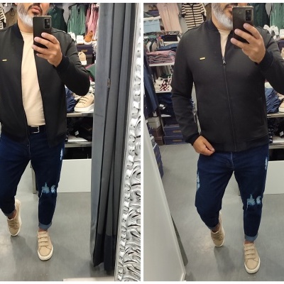 Homem a experimentar dois casacos pretos em provador com calças de ganga e sapatilhas bege