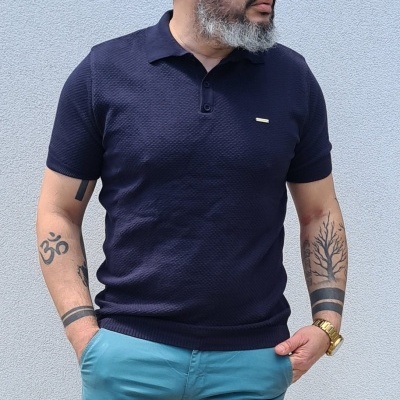 Homem com polo azul escuro texturizado e calças azul claro junto a uma parede cinzenta.
