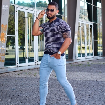 Homem com polo de riscas brancas e pretas, calças azul claro e óculos de sol em frente a edifício moderno.