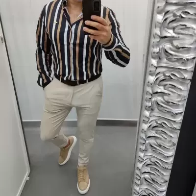 Homem com camisa às riscas, calças bege e sapatos desportivos perante espelho com moldura prata ondulada