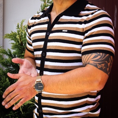Camisa de malha às riscas brancas, castanhas e pretas com botões, usada por homem com relógio e tatuagem, plantas e porta ao fundo