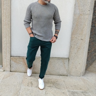 Homem com camisola cinzenta, calças verdes e sapatos brancos em ambiente urbano de pedra