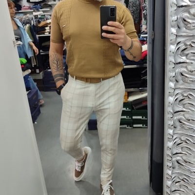 Homem com t-shirt castanha, calças bege xadrez e sapatos castanhos num espelho de loja de roupas