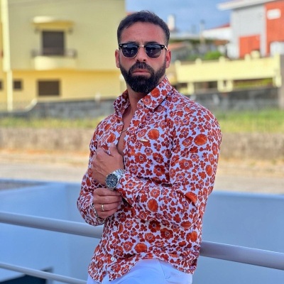 Homem com camisa floral laranja e branca, calças brancas e óculos escuros ao ar livre