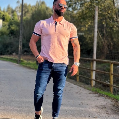 Homem a caminhar com polo rosa, calças de ganga e ténis pretos numa estrada com vegetação