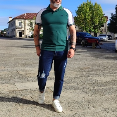 Homem com polo verde e branca, jeans rasgados e tênis brancos numa praça de pedra