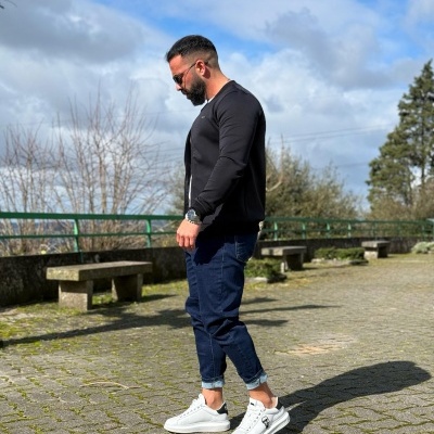 Homem com cardigan preto, calças azuis e sapatilhas brancas em calçada de pedra ao ar livre