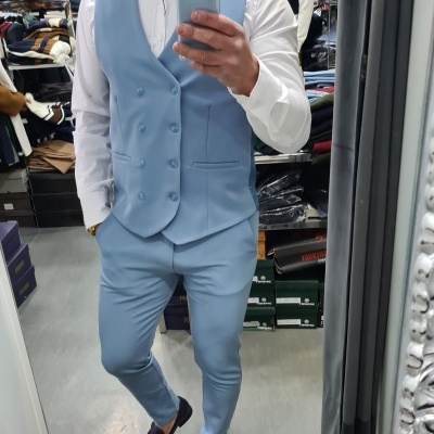 Homem com colete azul claro, calças azuis, camisa branca e sapatos pretos com laço vermelho num provador