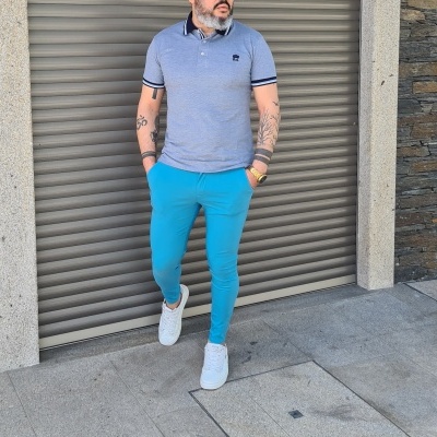 Homem vestido com polo azul claro, calças azuis e ténis brancos em calçada junto a porta metálica