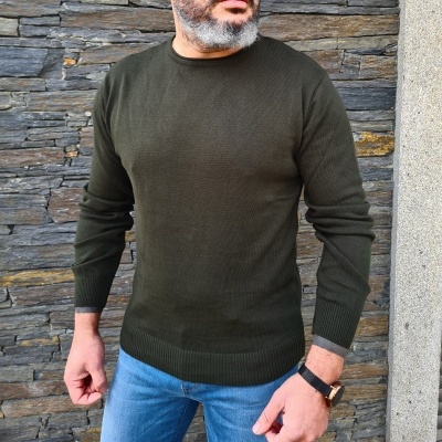 Homem a usar camisola verde escura de malha de gola redonda com parede de pedra ao fundo