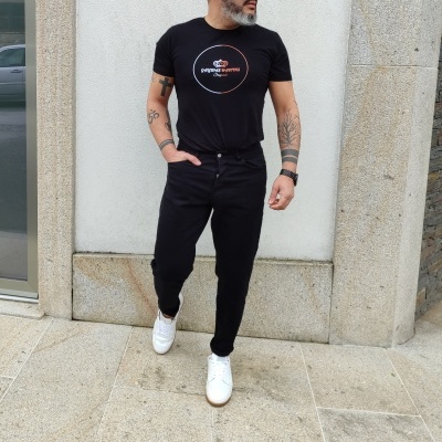 Homem com t-shirt preta com logo, calças pretas e sapatilhas brancas junto a parede de pedra clara