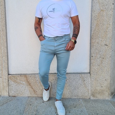 Homem vestido com t-shirt branca, calças azul claro e sapatilhas brancas junto a parede de pedra cinza