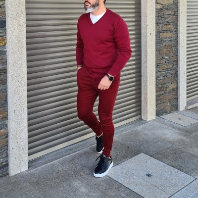 Homem a usar conjunto vermelho vinho com camisola de malha e calças, calçado desportivo preto, junto a portão metálico e parede de pedra.