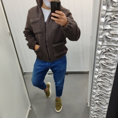 Homem a experimentar casaco castanho felpudo num provador com espelho decorado.