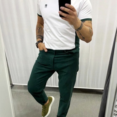 Homem com t-shirt branca e calças verdes a tirar selfie em espelho, fundo de cortinas brancas