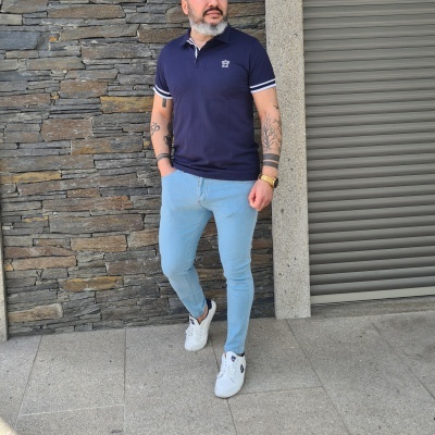 Homem com polo azul escuro, calças de ganga azuis claras e sapatilhas brancas numa rua com parede de pedra.