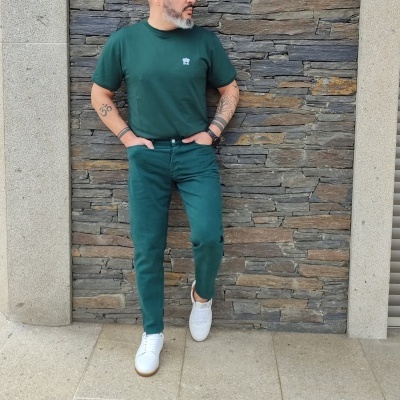 Homem com roupa verde escura encostado numa parede de pedra cinzenta