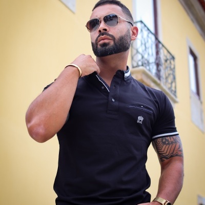 Homem a usar camisola polo preta com detalhes brancos e acessórios dourados em fundo de parede amarela