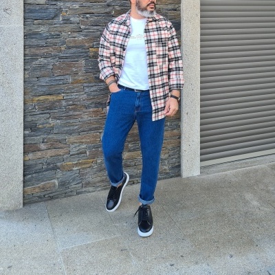Homem com camisa xadrez, t-shirt branca, calças de ganga e sapatilhas pretas em fundo urbano de pedra