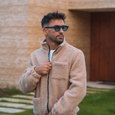 Homem com casaco felpudo bege e óculos escuros ao ar livre junto a parede de tijolos