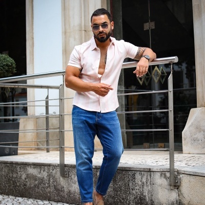 Homem de camisa rosa claro, calças de ganga azuis e sapatos castanhos ao ar livre encostado a corrimão metálico