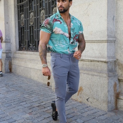 Homem com camisa estampada, calças cinza e sapatos pretos numa rua com calçada de pedra