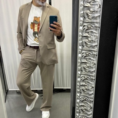 Homem com fato bege, camisa branca com urso estampado, cinto castanho e ténis brancos a tirar selfie num espelho prateado.