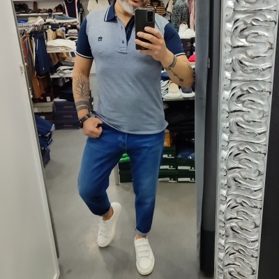 Homem com camisa polo cinza, jeans azuis e sapatilhas brancas num espelho com moldura prateada