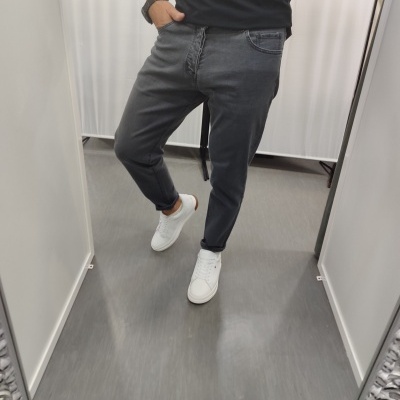 Pessoa a vestir calças de ganga cinza escuras, sapatilhas brancas e t-shirt preta, tirada em frente a espelho de provador