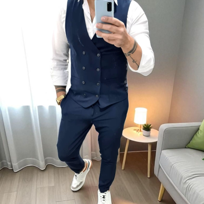 Homem com colete e calças azul escuro, camisa branca e ténis brancos, de pé numa sala com sofá cinzento e almofada verde