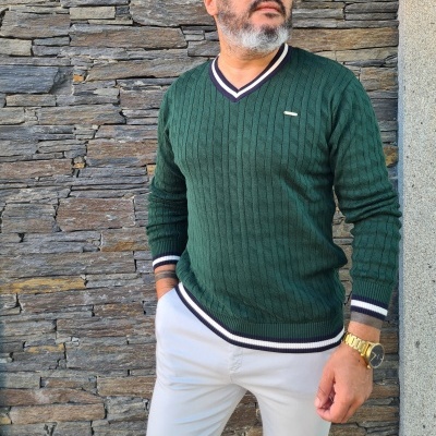 Homem com camisola verde escura e calças brancas em frente a parede de pedra