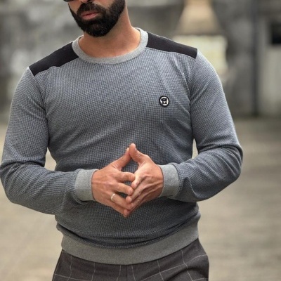 Homem vestido com camisola cinzenta e calças cinzentas xadrez, com óculos de sol pretos, ao ar livre.