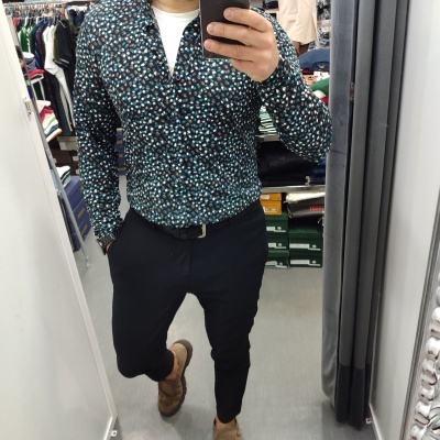 Homem com camisa de padrão de pontos coloridos, calças pretas e sapatos castanhos num espelho de loja de roupa