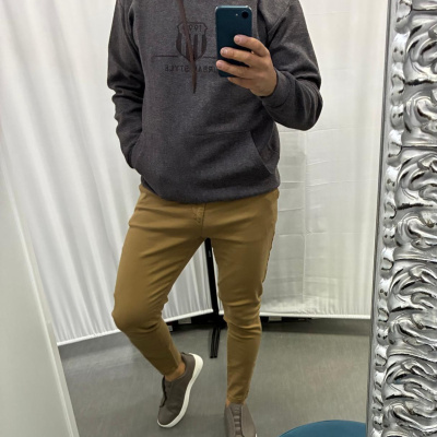 Homem de pé num provador a usar sweatshirt castanha escura com capuz e calças castanhas claras, a tirar selfie num espelho decorativo.