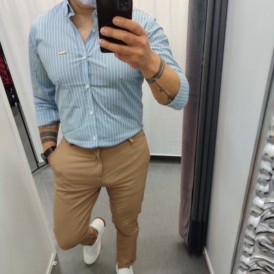 Homem com camisa azul riscada, calças camel e sapatilhas brancas em provador